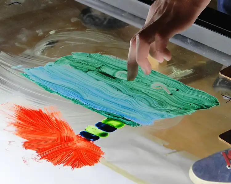 Finger verwischt grüne und orange Farbe auf einer Glasplatte.