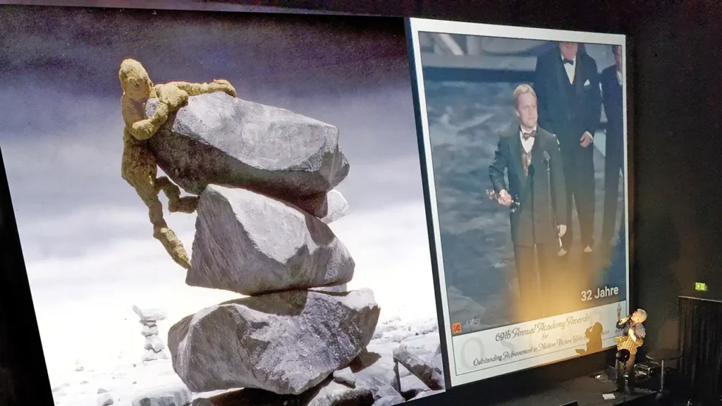 Thomas Stellmach in front of the screen on which he shows a picture, one of the Oscar-winning film QUEST and one of himself holding the Oscar as he gives his speech on stage at the awards ceremony in Hollywood.