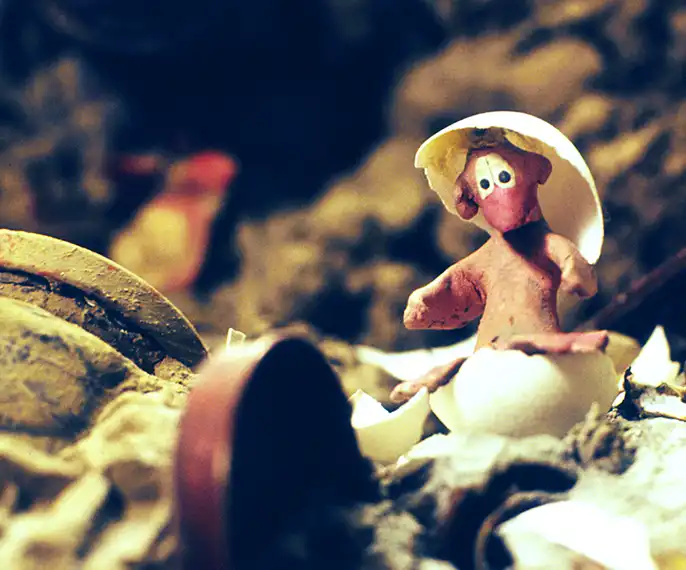 Film still from LET ME ALSO LIVE. The image shows the main character, a chicken made of plasticine that has just hatched, sitting in half an eggshell and wearing the top half of the shell on its head.